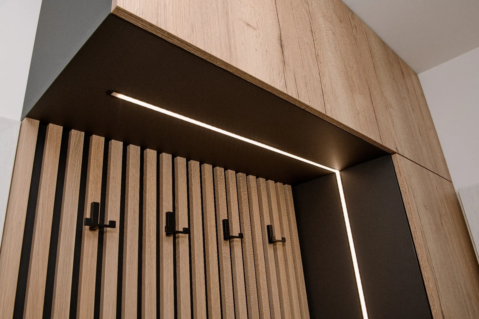 Předsíňová stěna s lamelami a LED Vestavná předsíňová stěna s dubovými lamelami, černými háčky a integrovaným LED osvětlením.